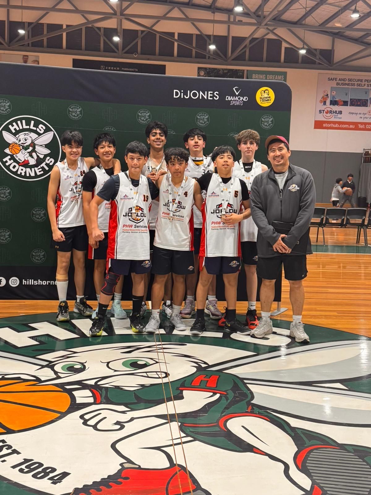 Youth basketball team posing on court, wearing red and white jerseys. Green backdrop.