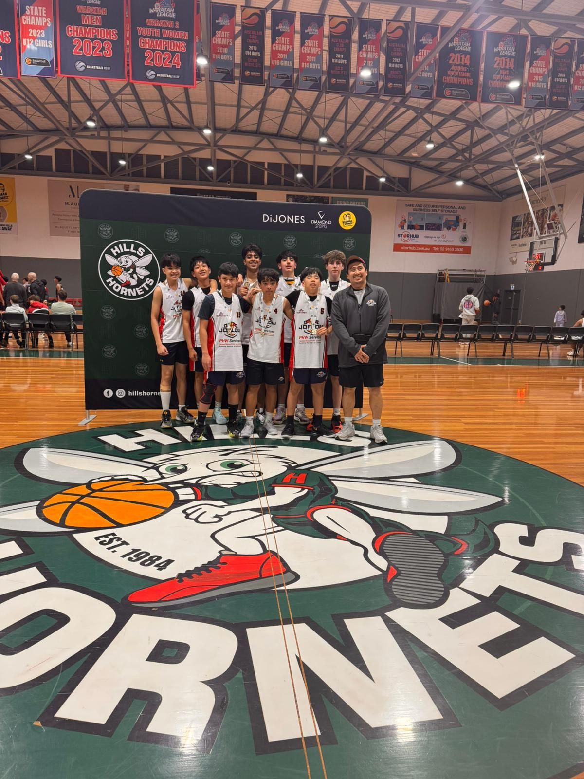 Basketball team posing on a Hornets court, in front of a banner.