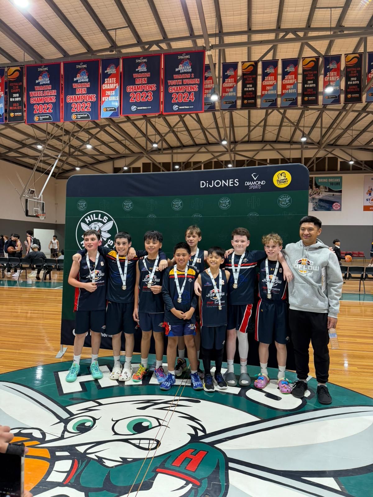 Basketball team, wearing medals, poses on a court after a tournament. They stand with a coach in front of a banner.