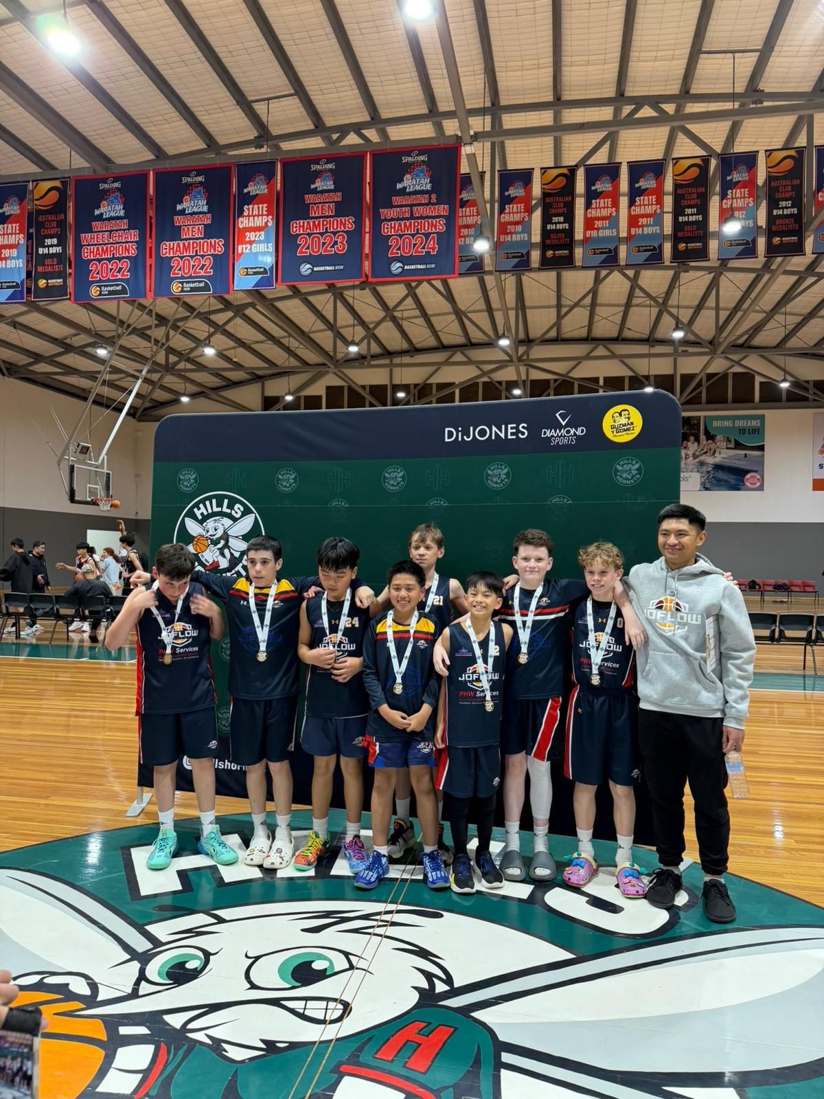 Basketball team posing for a photo on a podium, wearing medals. Indoors, green and white bunny logo on the floor.