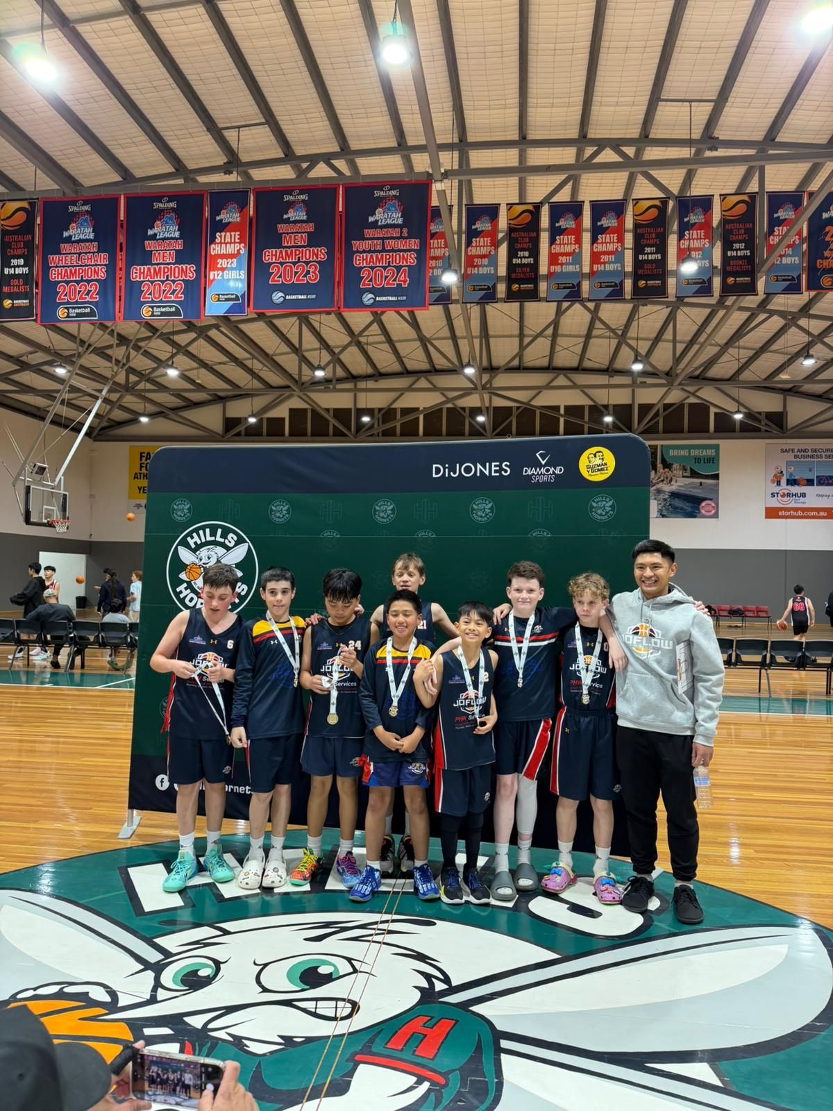 Youth basketball team posing on a green mat, each wearing a medal. Indoor court with banners above.