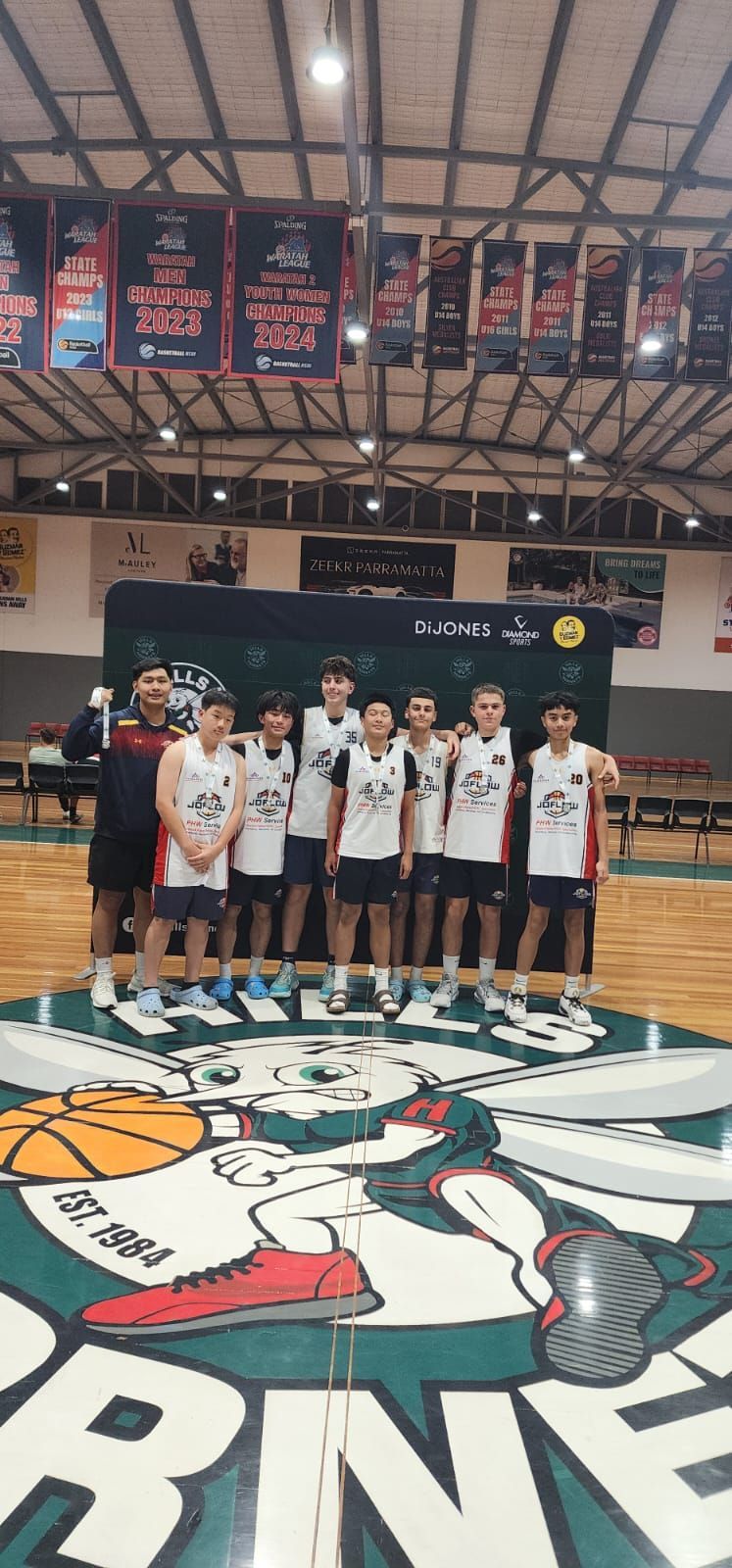 Basketball team posing on a court with a logo. Green, white, and black design; banners overhead.