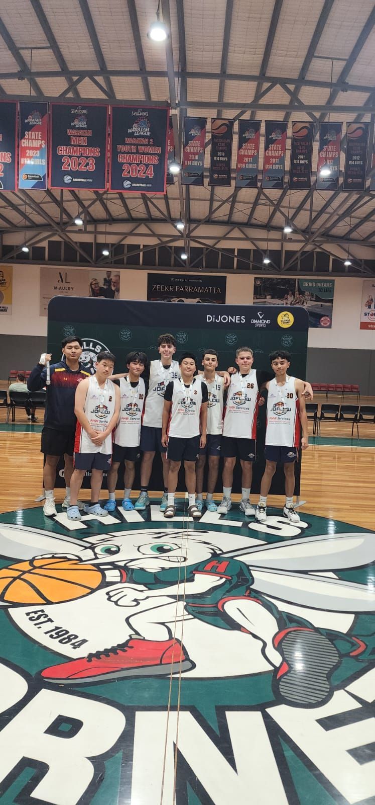 Basketball team poses on a Hornets logo court. Players in white jerseys, black shorts, and sneakers stand with coach.