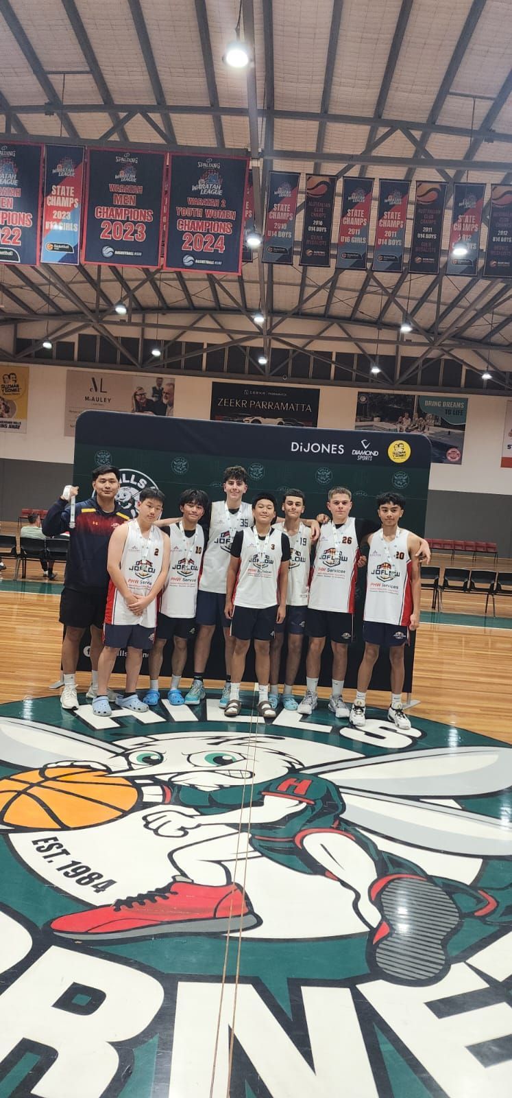 Basketball team posing on a court, team is wearing jerseys, with a basketball logo in the floor.