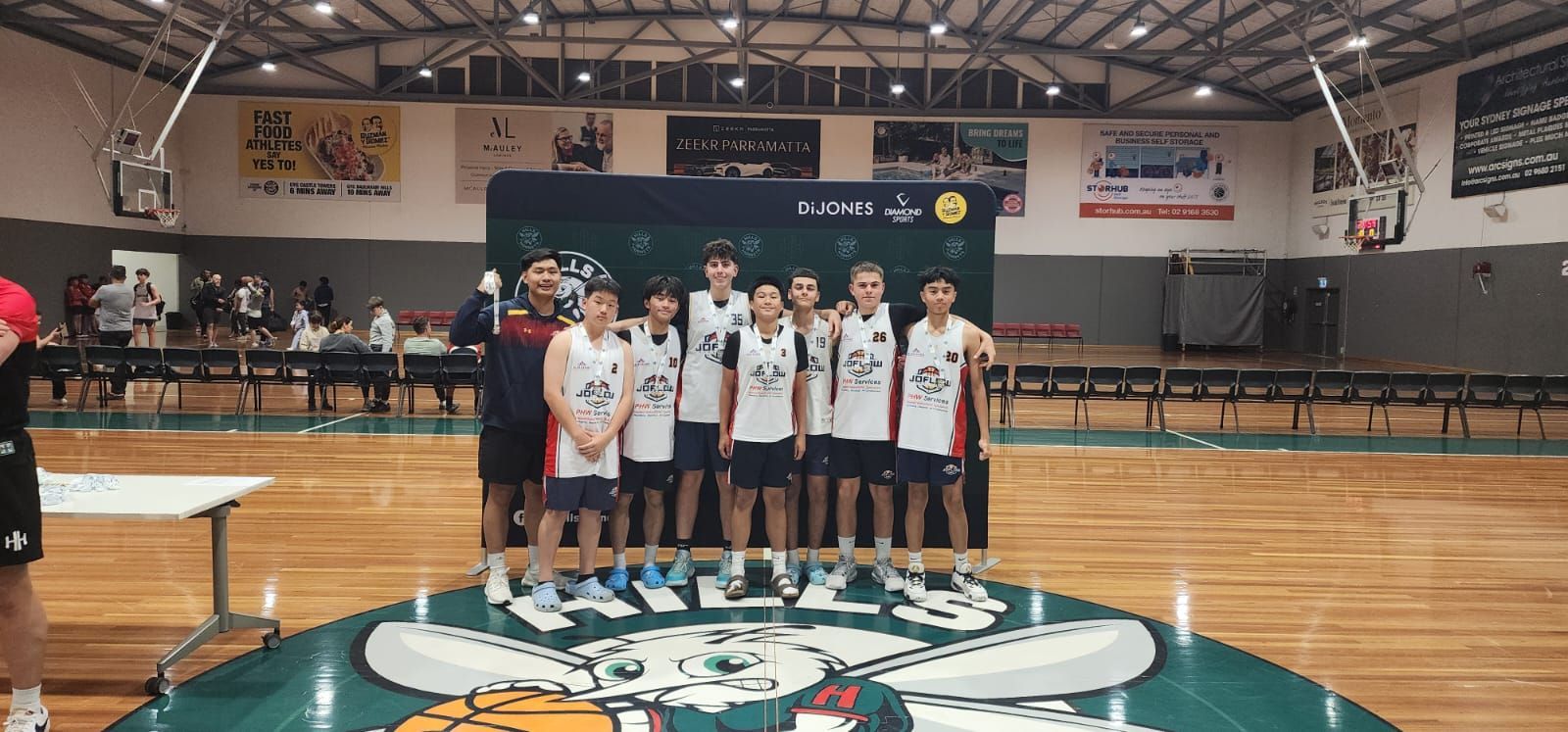 A basketball team poses on the court. They are wearing jerseys. Court is made of wood.