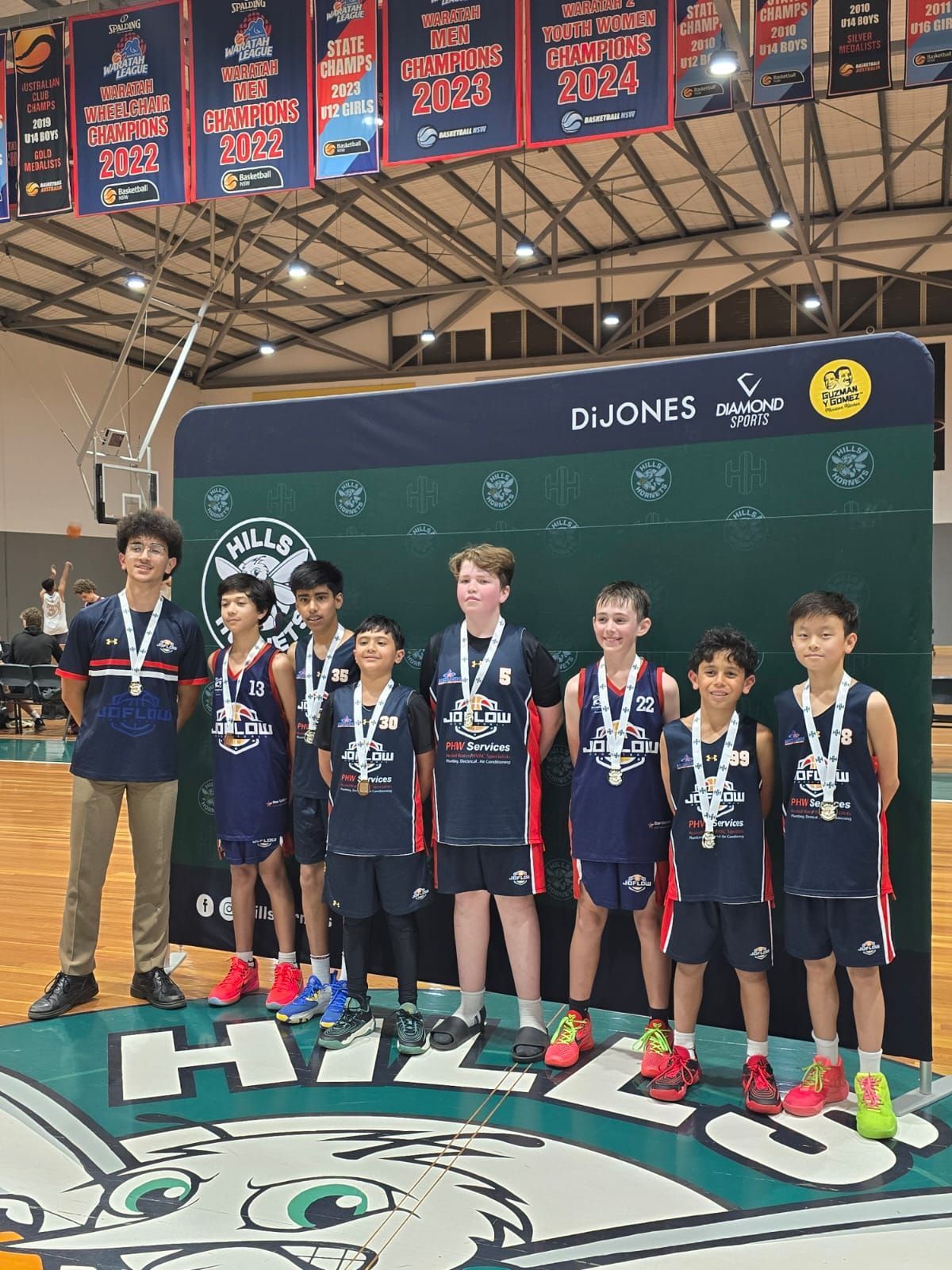 Youth basketball team on podium, wearing medals, celebrating a win. Indoors, team in dark blue uniforms.