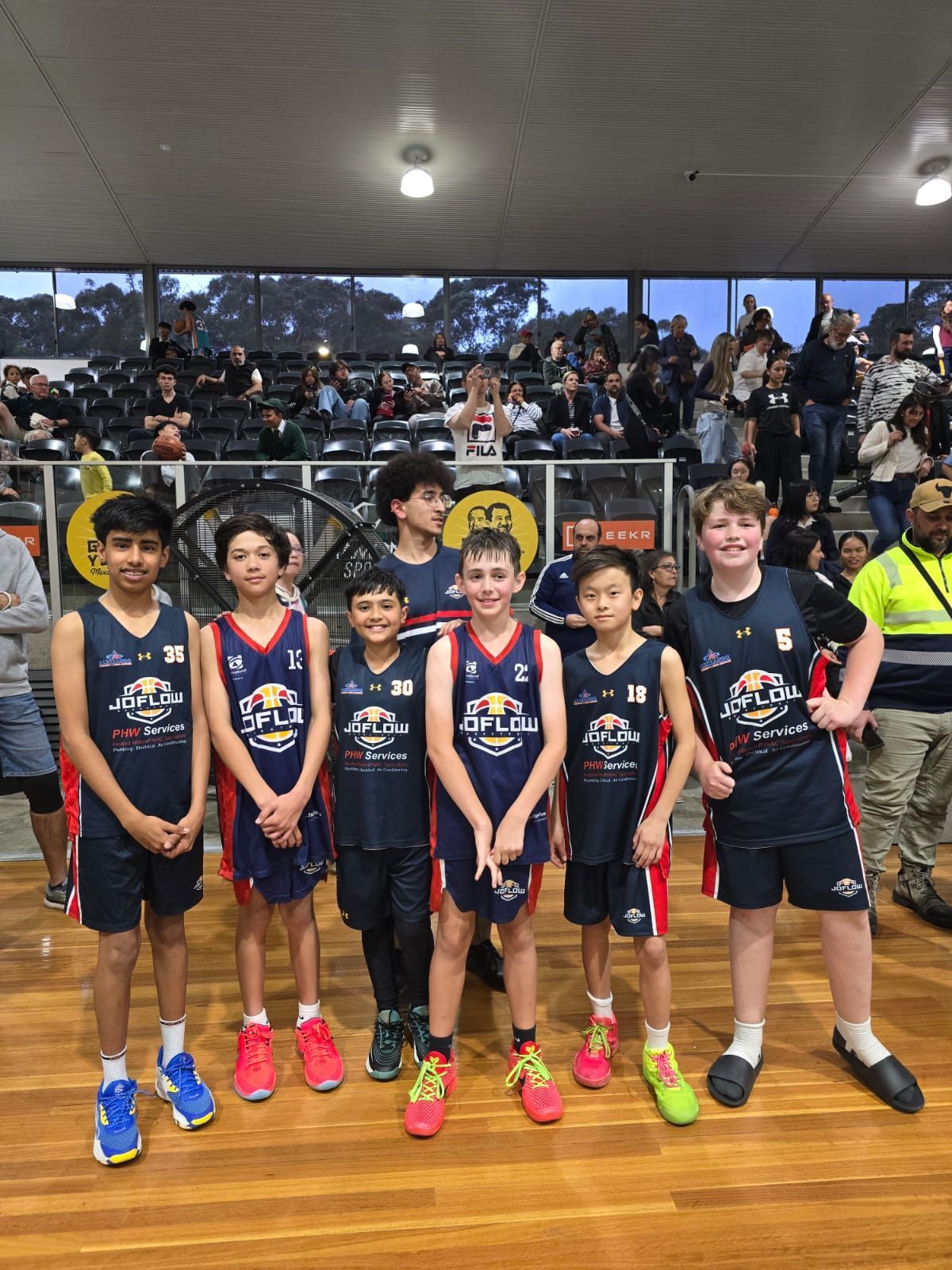 Youth basketball team poses on court, wearing navy jerseys with red and white accents. Crowd in background.