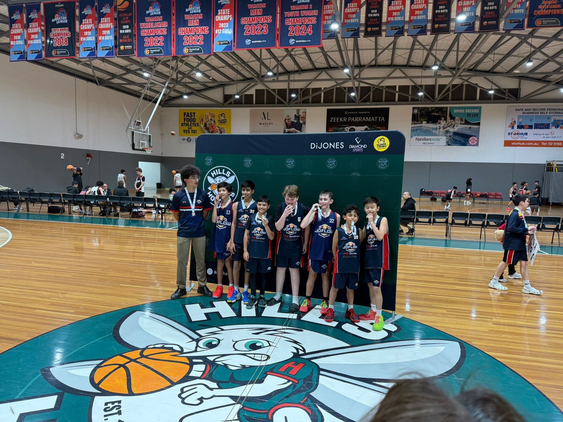 Youth basketball team on a podium after a game, each wearing a medal. The setting is a gymnasium.