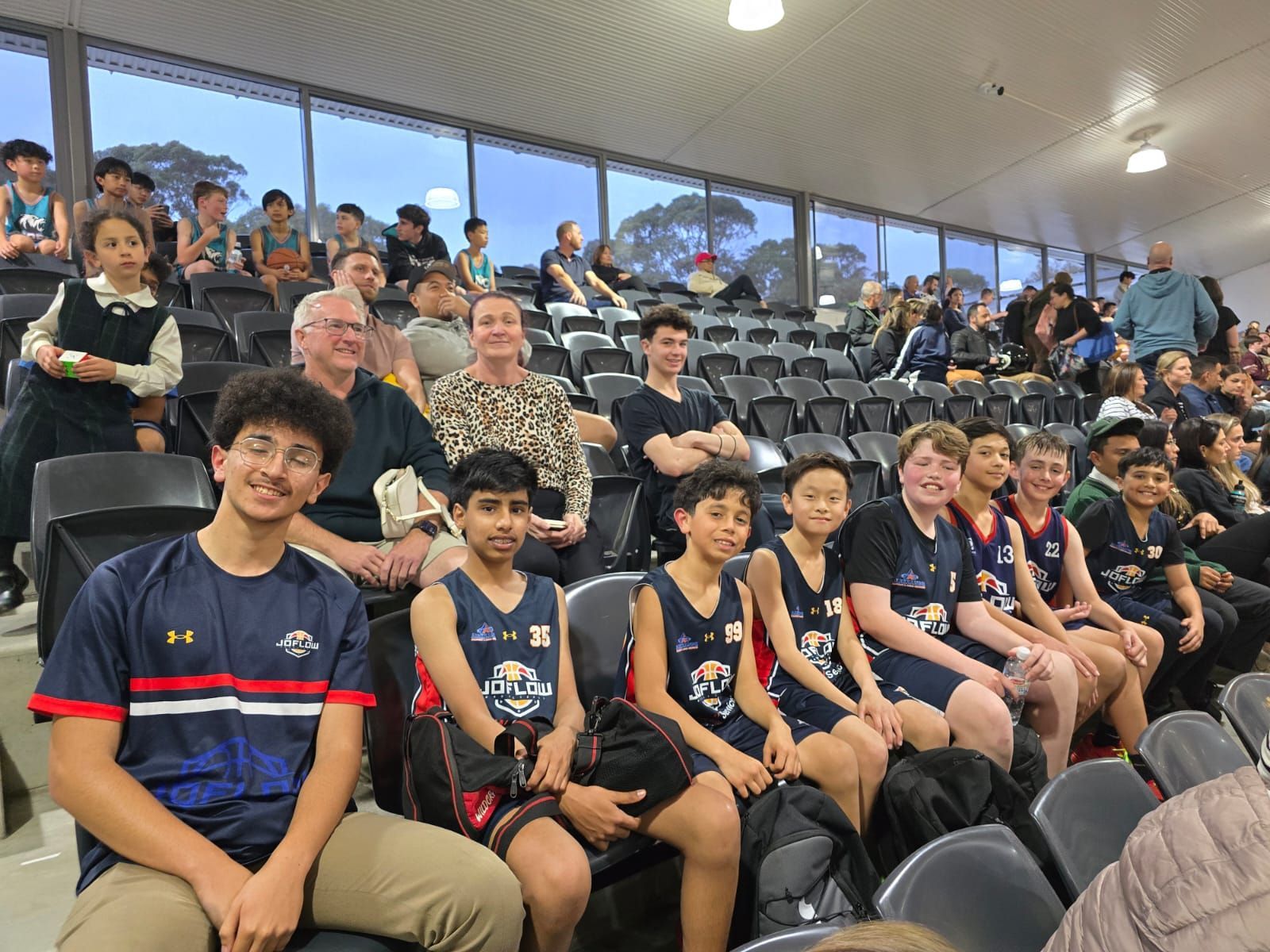 Basketball game: Crowd watching, some players in uniform seated in foreground, others in background, indoor setting.