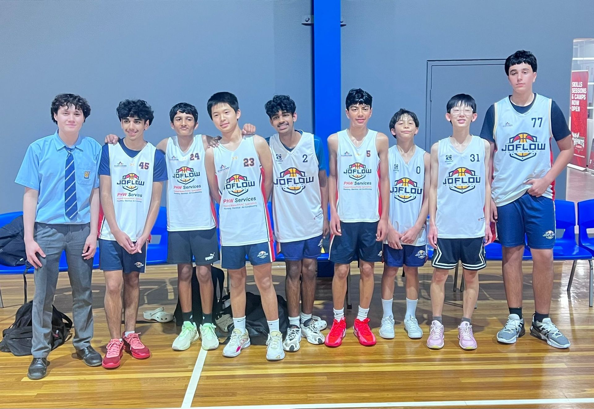 Basketball team posing indoors, wearing white jerseys and blue shorts. Several are smiling.