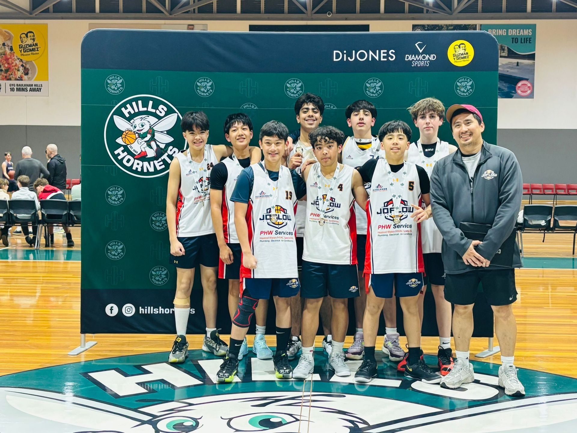 Basketball team, Hills Hornets, poses on court with coach and banner. Players in uniforms, green and white court.