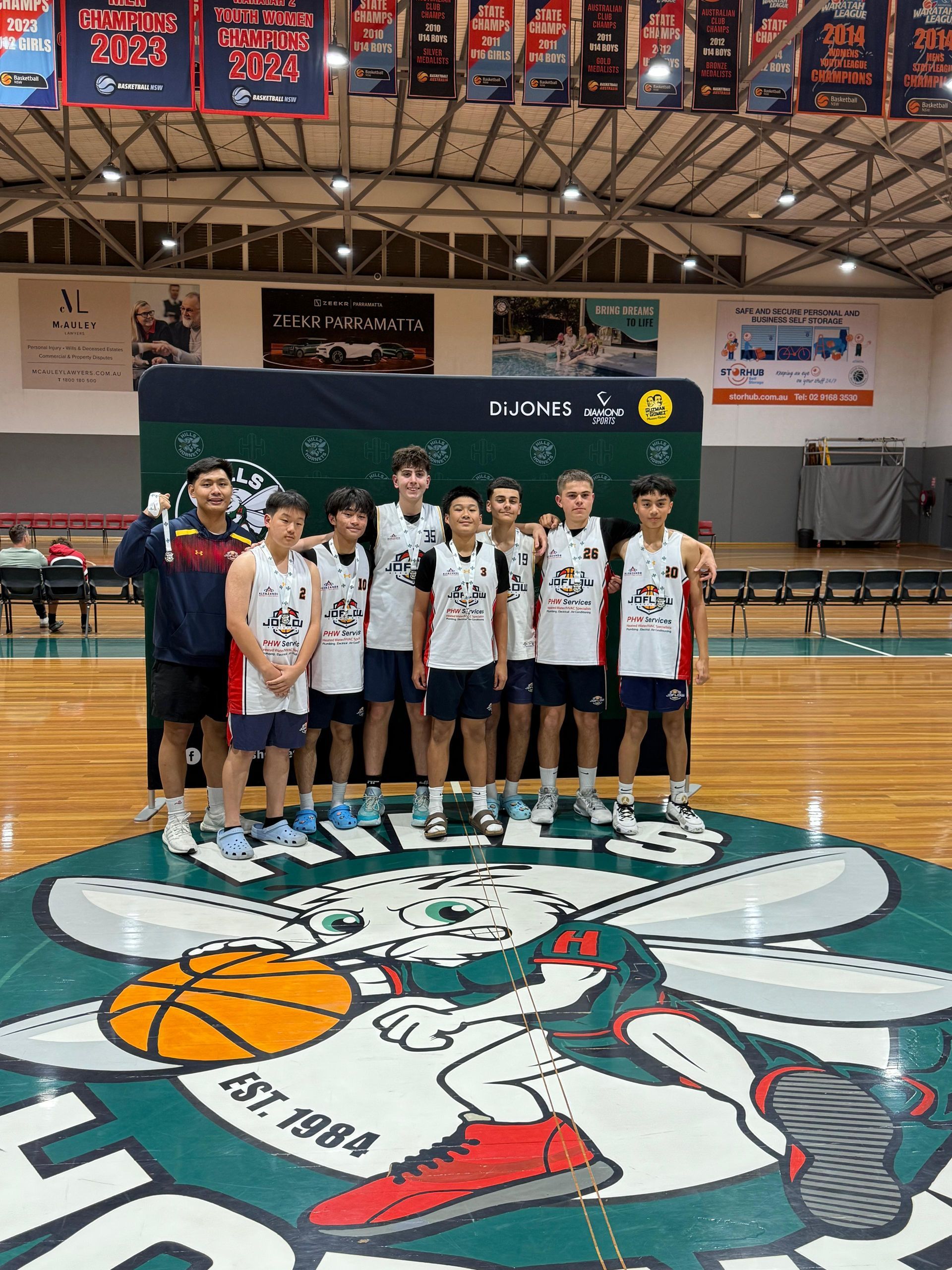Basketball team poses on court; players in uniforms with coach, indoor setting.