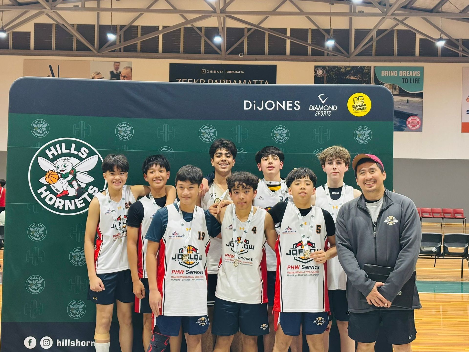 Basketball team, the Hills Hornets, pose in front of a banner, indoors. Players and coach smile, wearing Hornets jerseys.