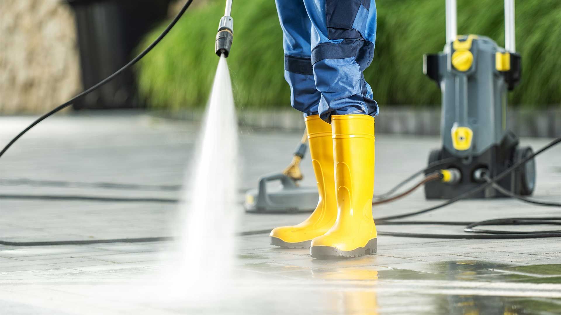 A person wearing yellow boots is using a high pressure washer on a sidewalk.