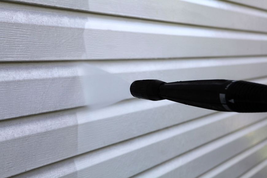 A person is cleaning a white siding with a high pressure washer.