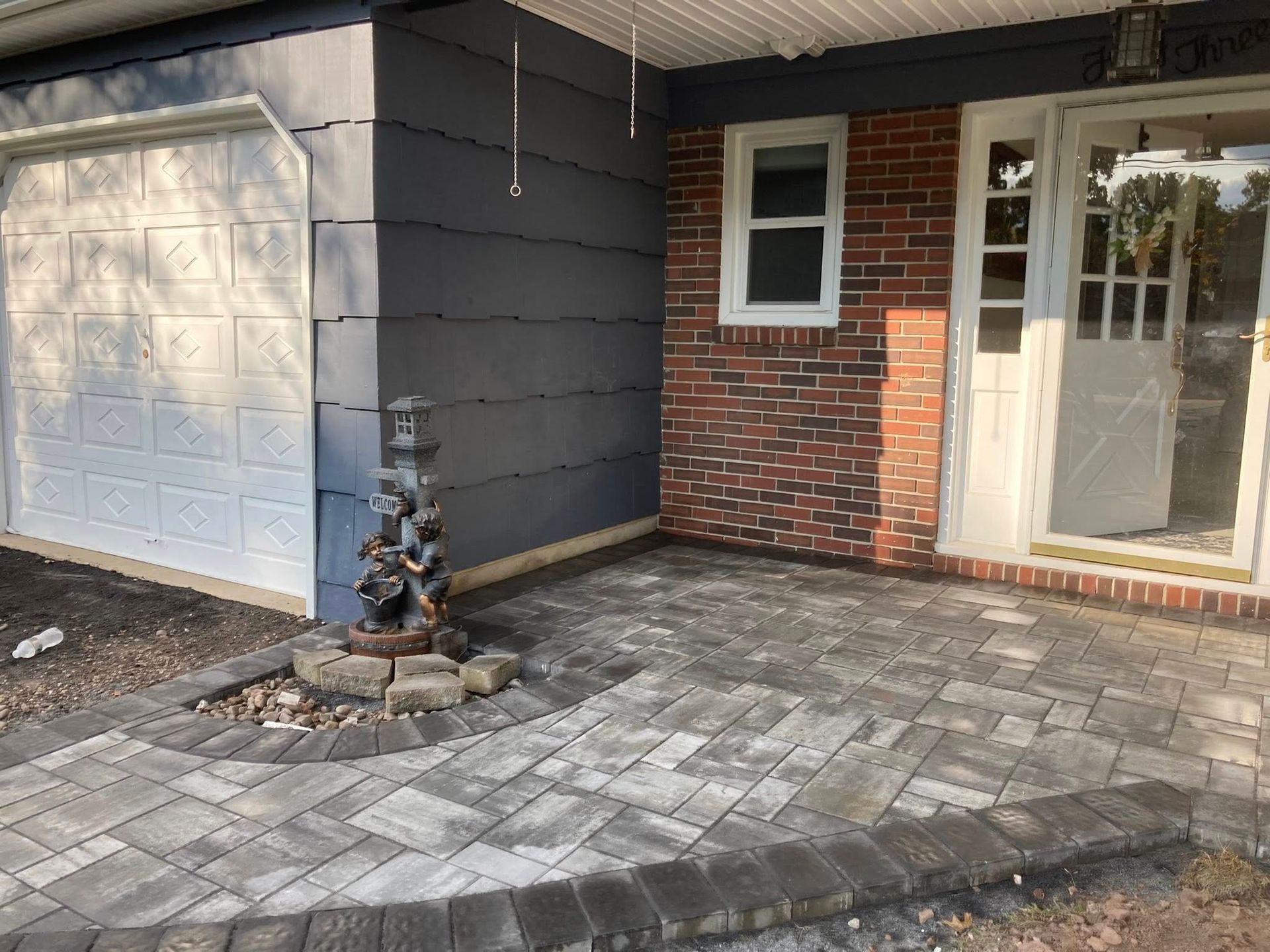 Entrance with a brick wall, paved walkway, fountain, garage door, and a white front door.