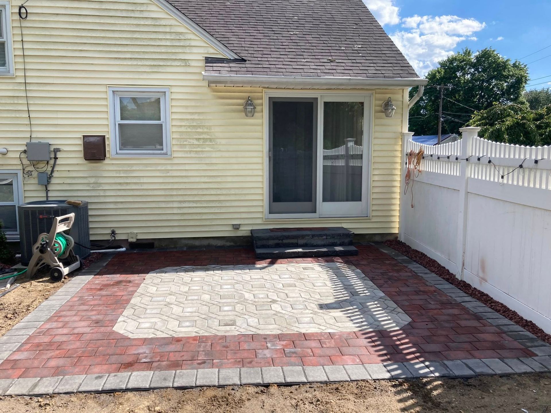 Backyard patio with red and tan pavers, next to a yellow house with a sliding glass door and white fence.