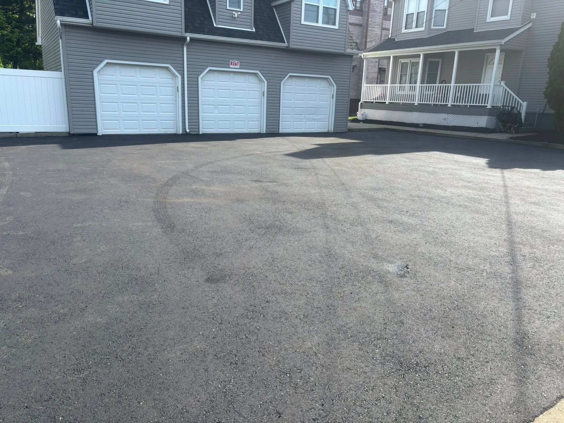 Asphalt driveway with three-door garage and a house with a porch in the background.