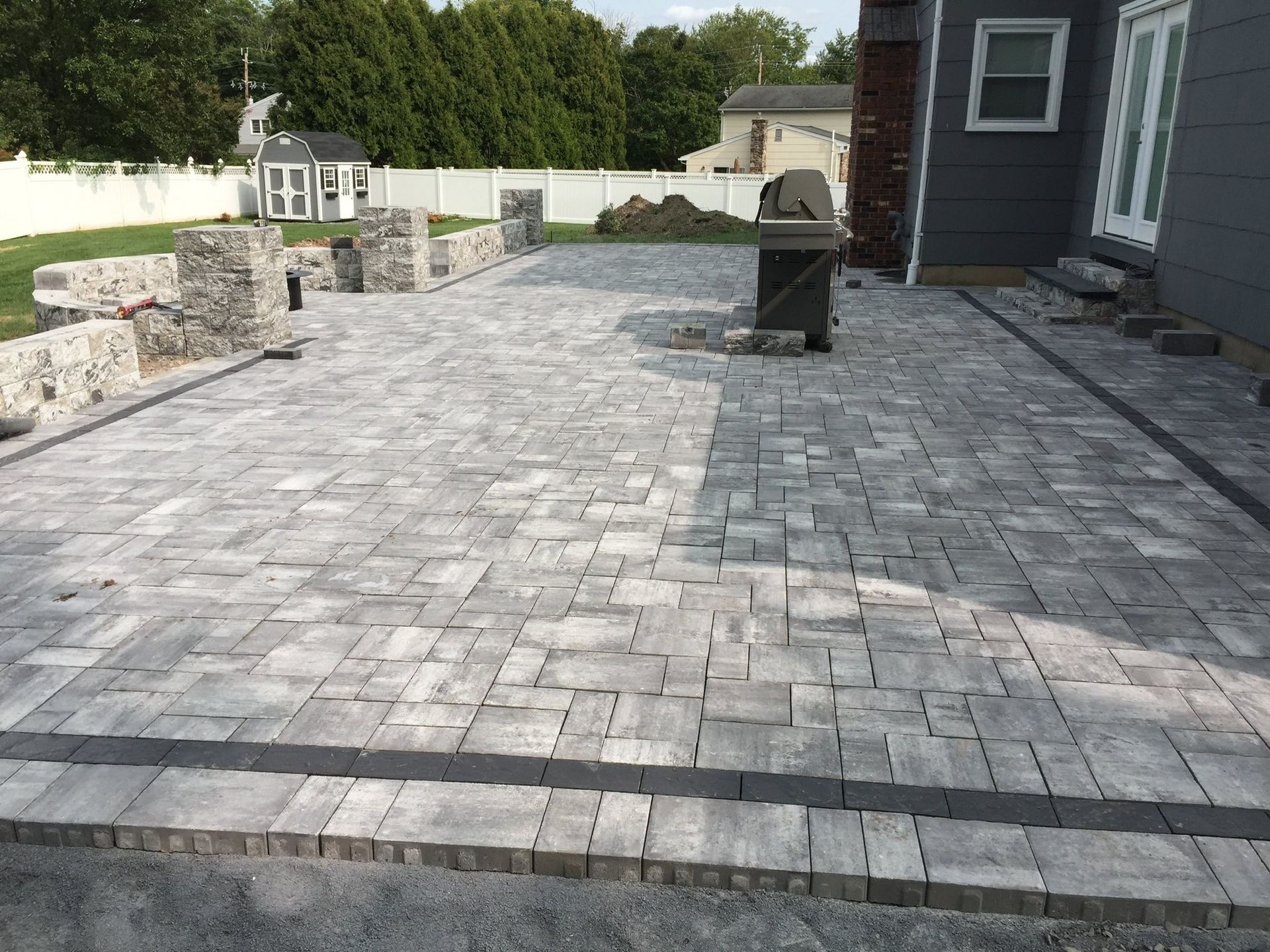 Gray paver patio with a dark border next to a house with a brick chimney and retaining walls.