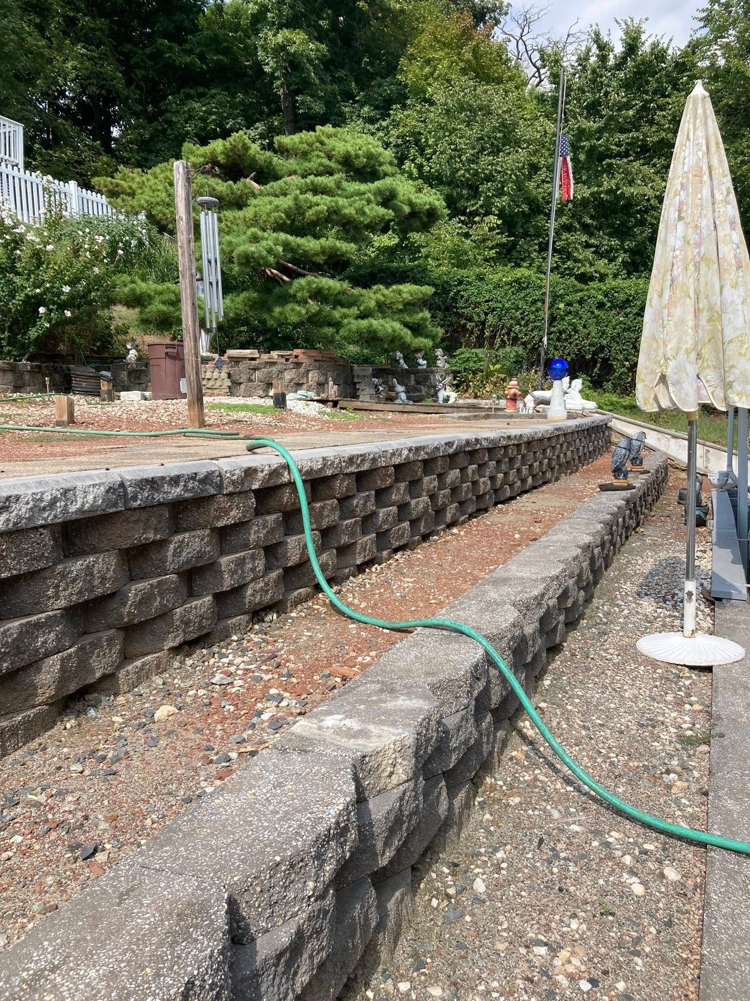 Long, stone wall with a green hose, next to a gravel path. Trees and an American flag are in the background.