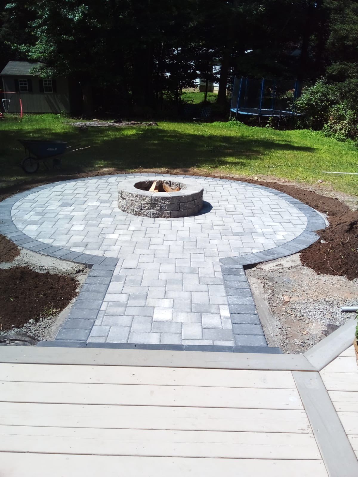 Circular paved patio with fire pit, pathway to wooden deck, surrounded by grass and trees.