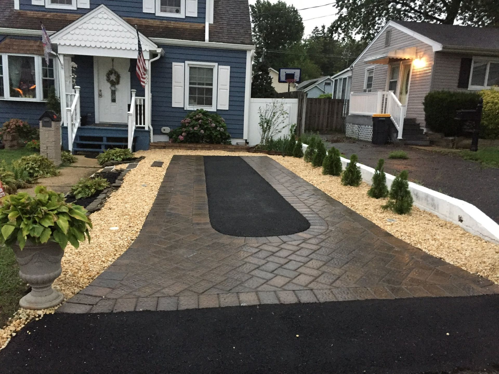 Driveway with brick pavers and asphalt. Blue house with a front porch. Gravel landscaping borders.