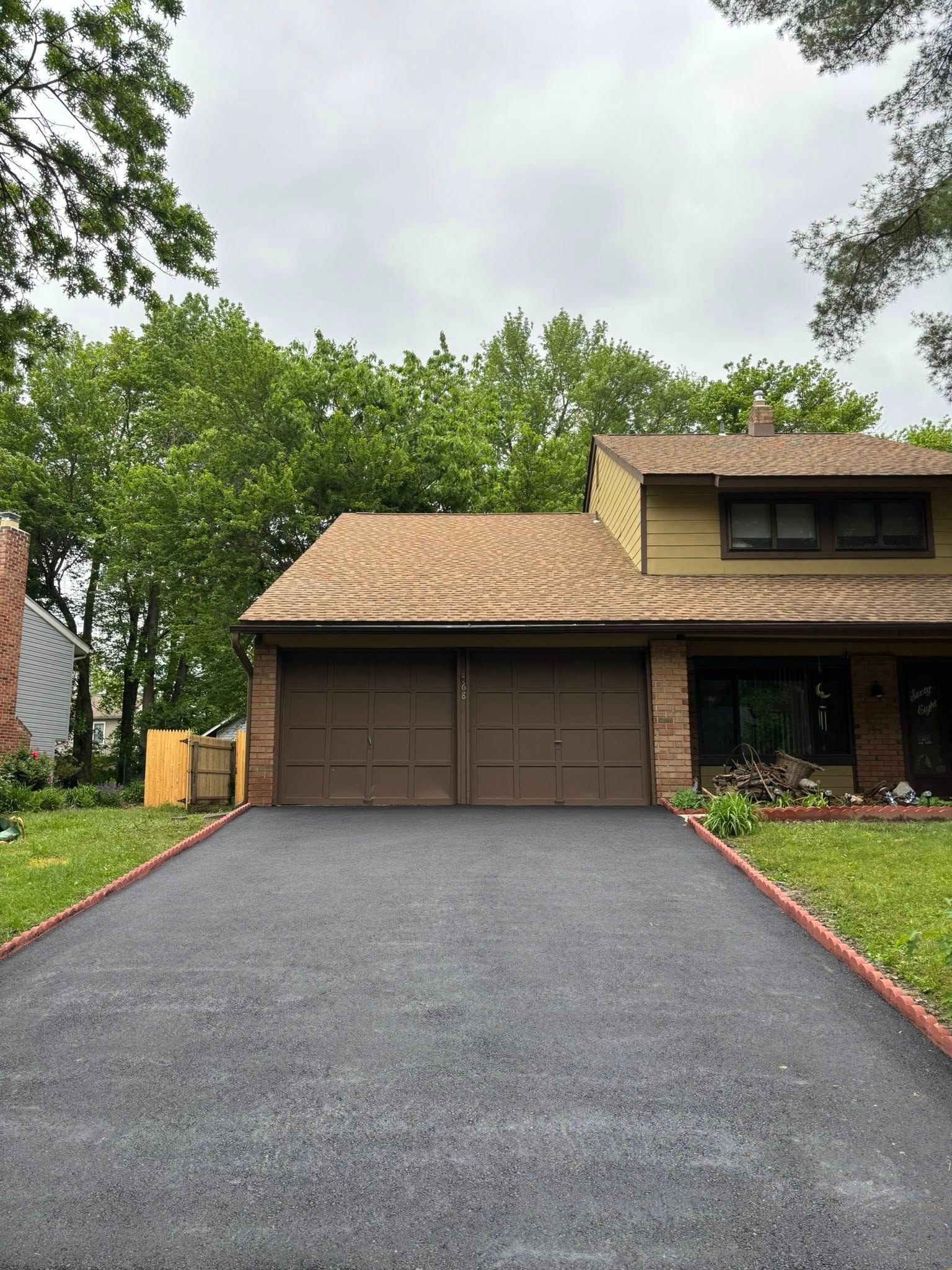Two-car garage with brown doors, a dark asphalt driveway, and green trees. The house has a brown roof and a tan exterior.
