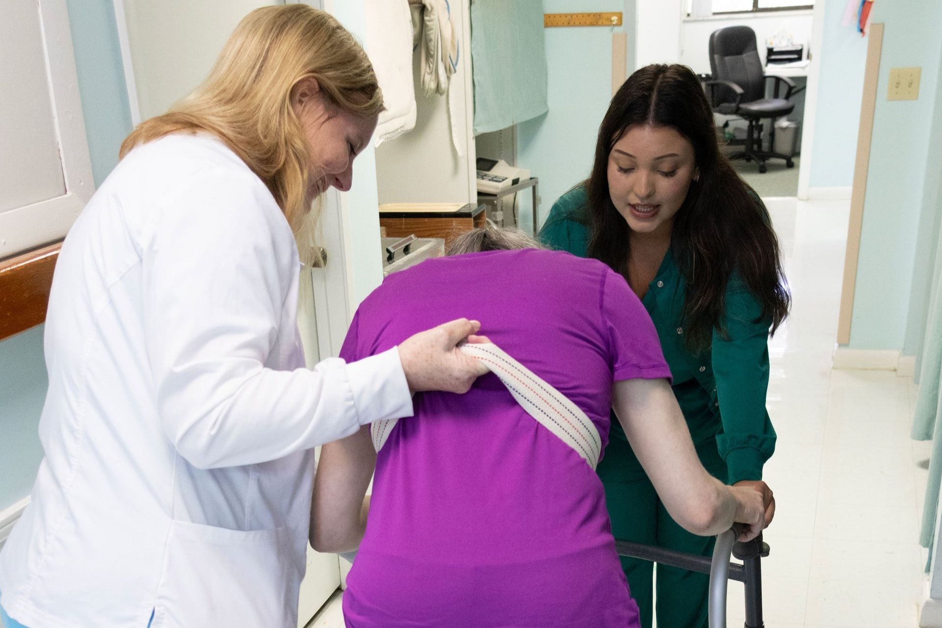 Two nurses are helping a patient with a walker in a hospital hallway.
