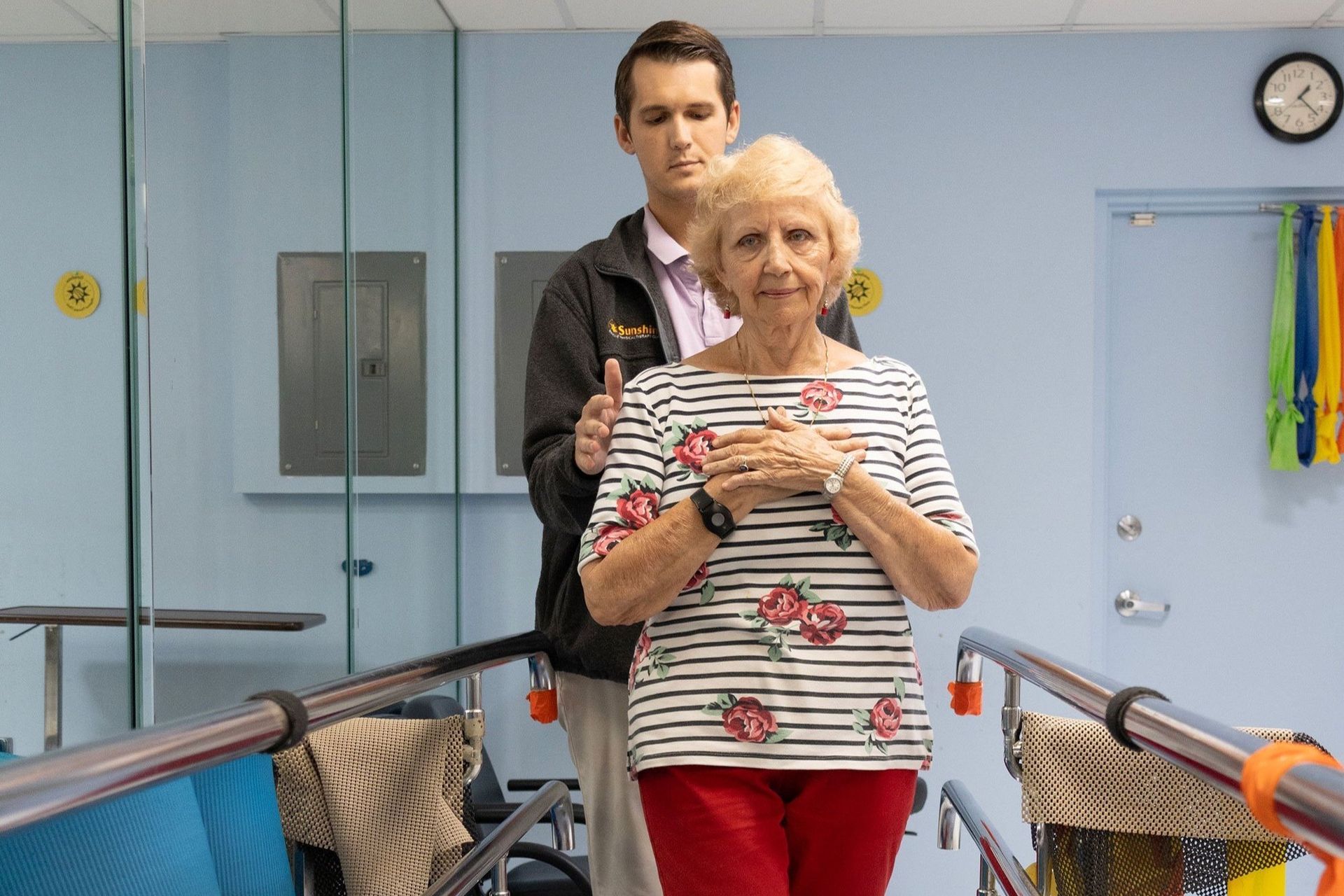 A man is helping an elderly woman walk on parallel bars.