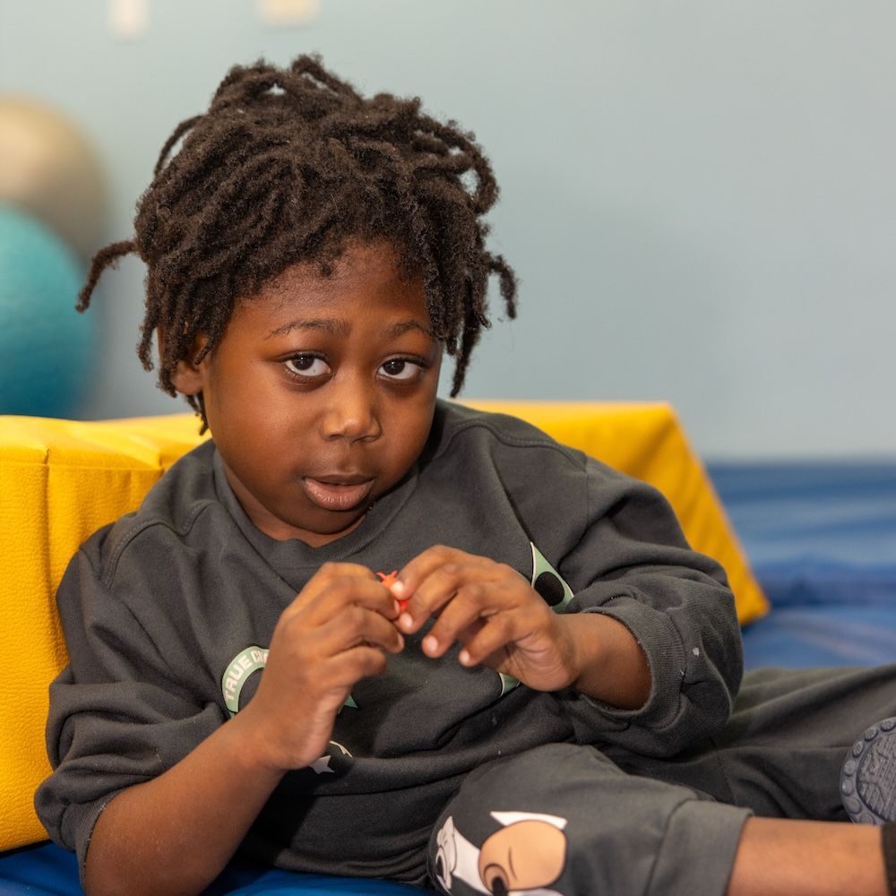 A child with dreadlocks in a grey sweatshirt sits on a blue and yellow foam mat, holding a small object in their hands.