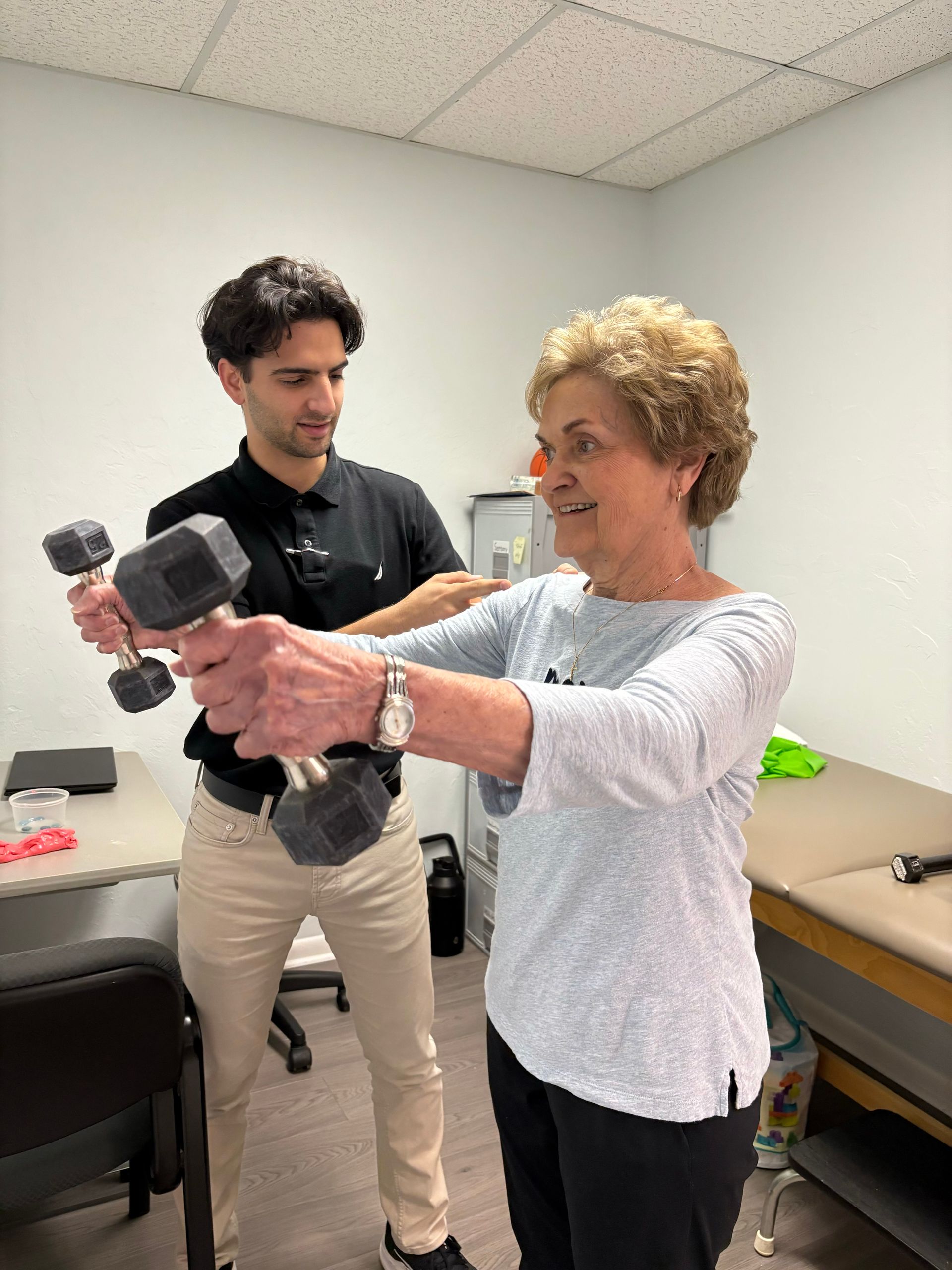 A man is helping an older woman lift a dumbbell.