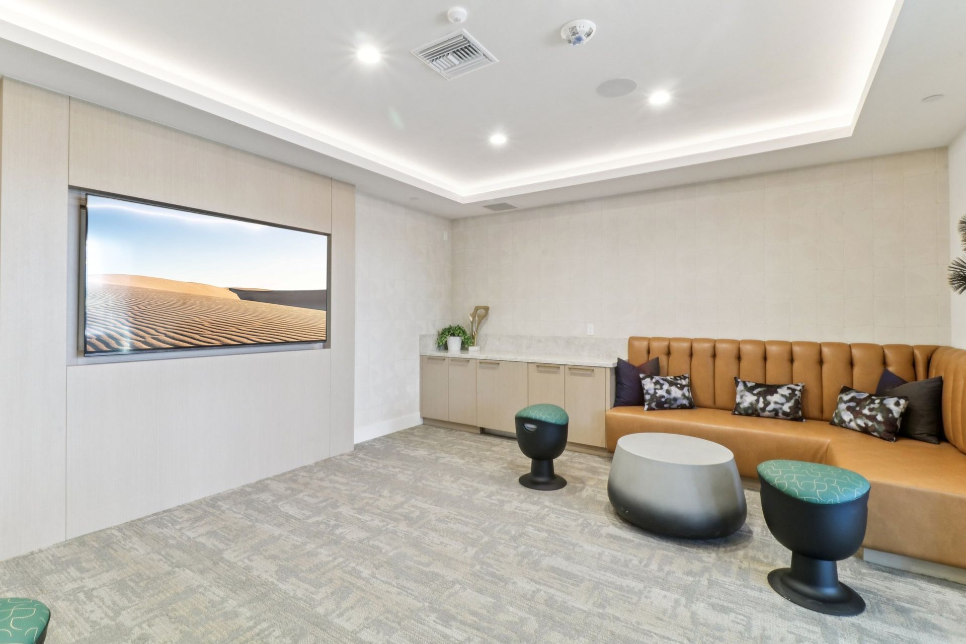 Lounge area with a large-screen TV, tan sectional sofa with throw pillows, and two small stools.