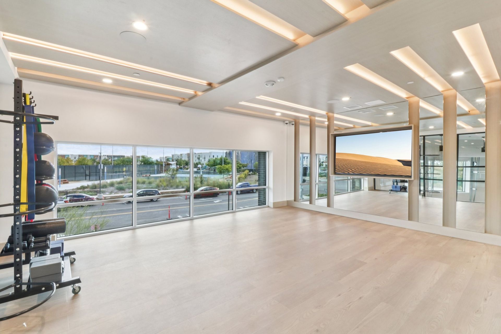 Bright, empty gym with large windows overlooking a street. Workout equipment is on the left; a large screen is on the right.