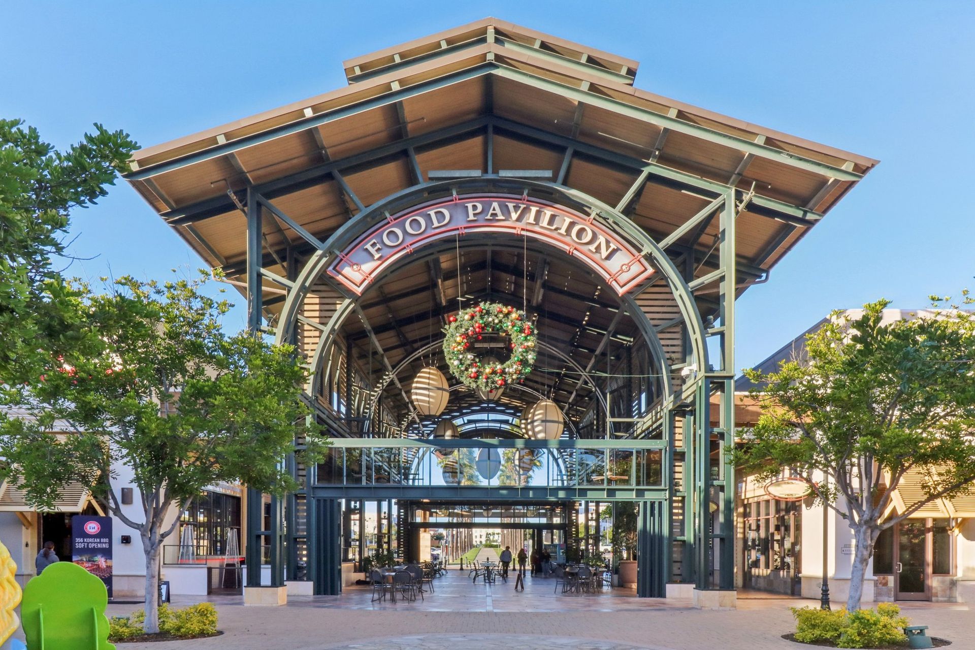Rooftop pavilion entrance with arched sign, metal framework, and storefronts; people walk beneath.