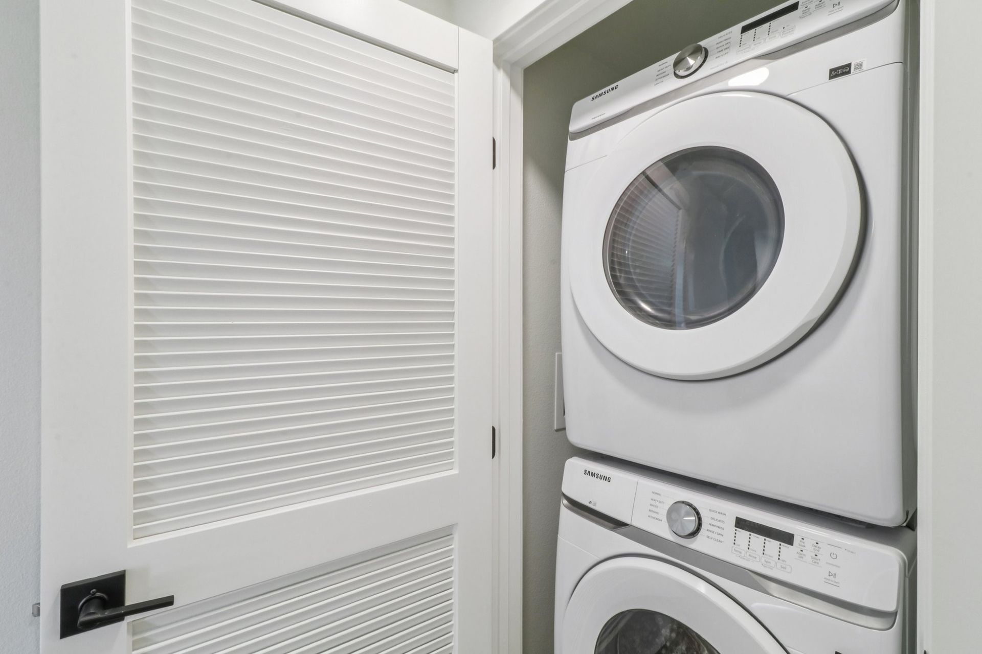 White stacked washer and dryer in a small laundry room. A white door is on the left.