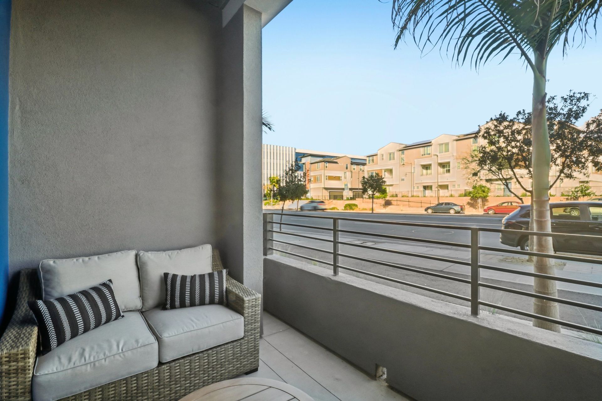Balcony with a wicker sofa and pillows overlooking a street with buildings and a palm tree.