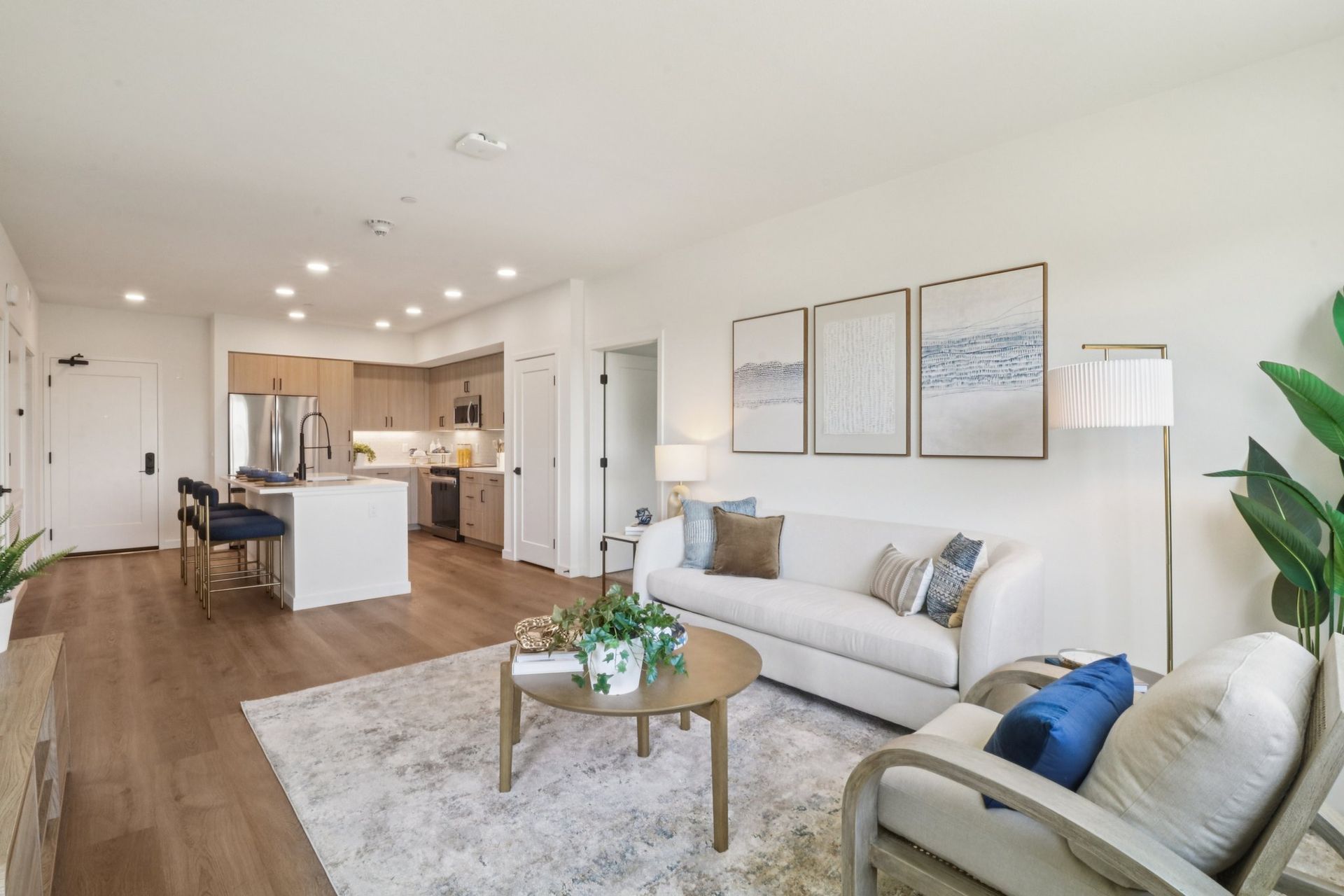 Open-plan living space: white sofa, framed art, kitchen with island, wood floors, and neutral tones.
