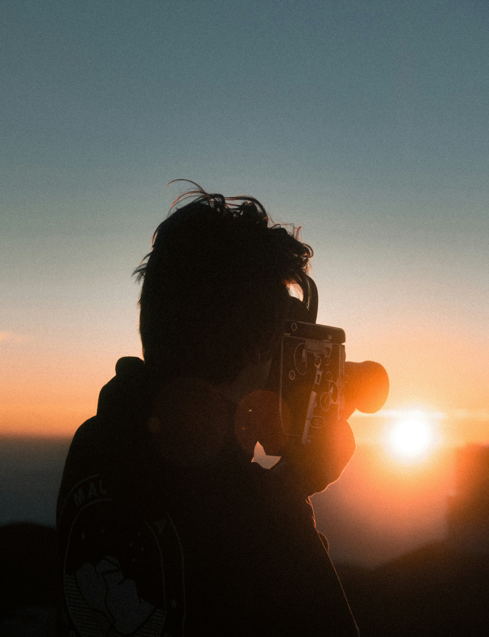 Silhouette of person taking photo of sunset with orange and blue sky.