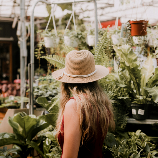 A woman wearing a hat is standing in a greenhouse surrounded by plants.