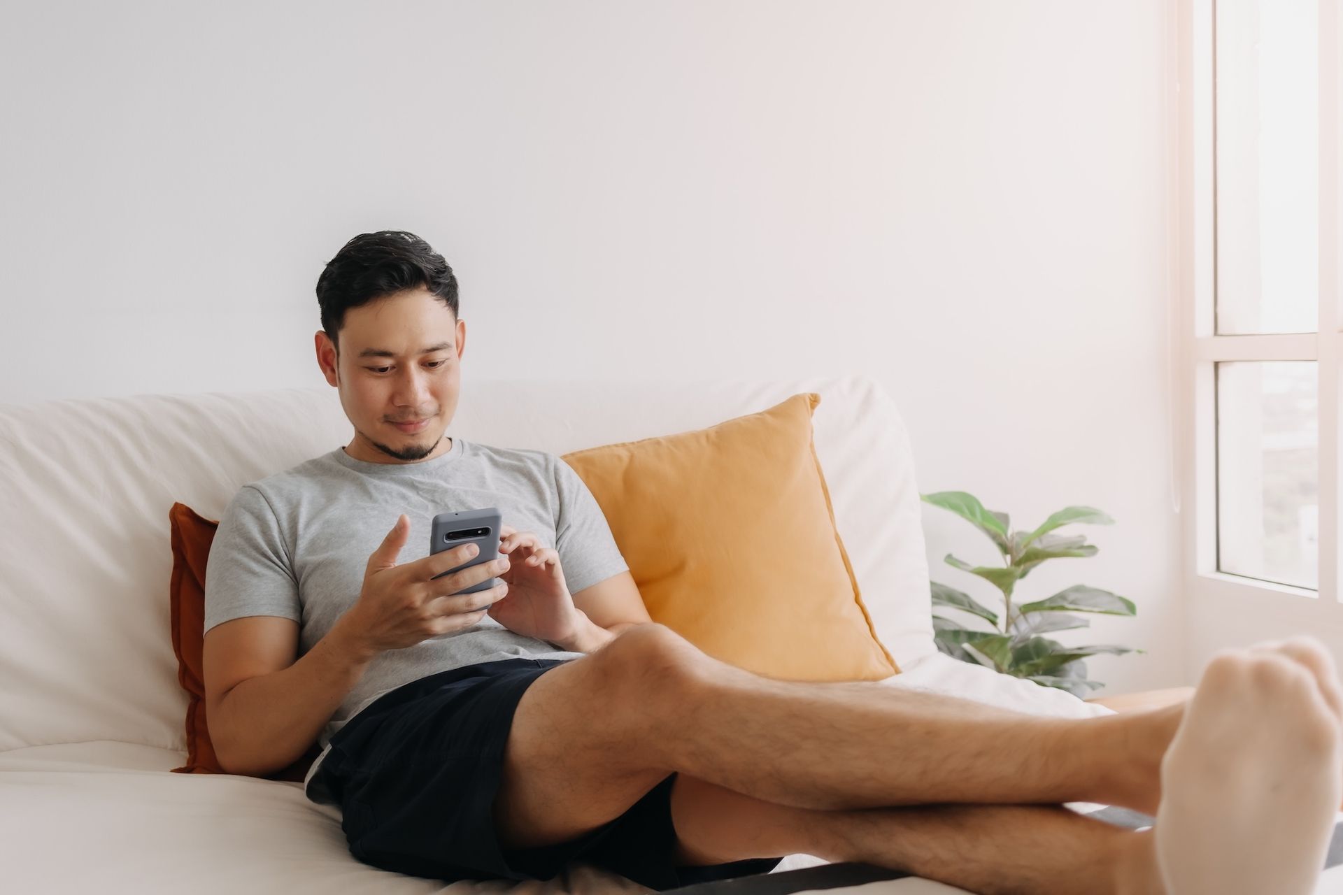 Man on a couch looking at a smartphone, relaxed in a bright room.