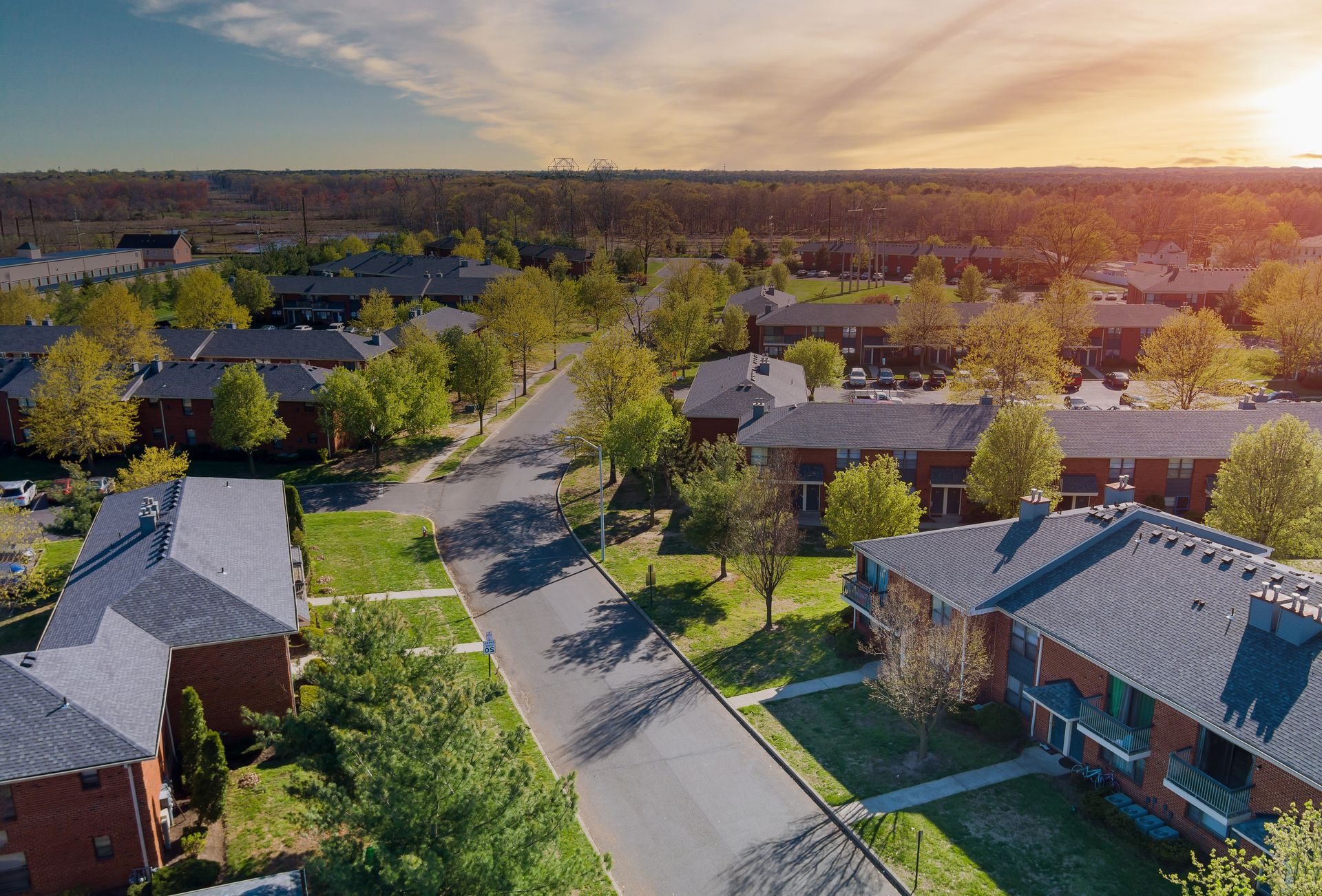 Aerial view of a suburban neighborhood with brick houses, green lawns, and a road under a sunny sky.