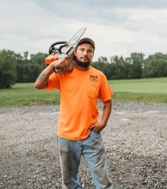 Man in orange shirt holds chainsaw on shoulder, standing outside on gravel, overcast sky.