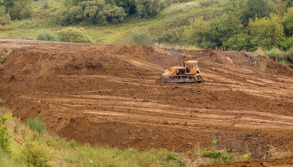 Bulldozer leveling a large pile of brown earth on a construction site; green foliage in the background.