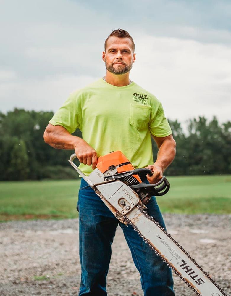 Man in a neon shirt holding a chainsaw, standing outdoors in front of a field and overcast sky.