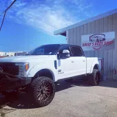 White Ford truck with large wheels parked outside David's Body Shop, sunny day.