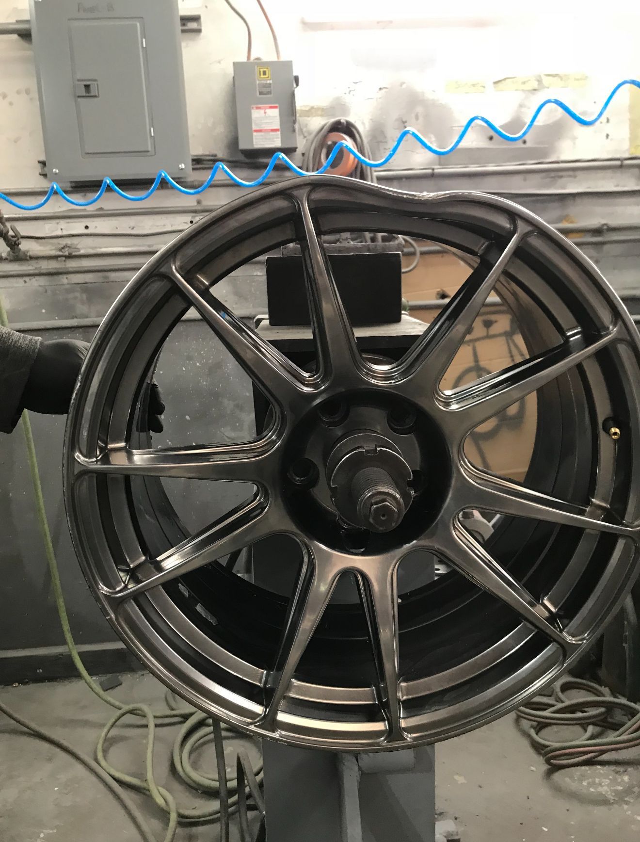Bent, polished alloy car wheel on a stand in a workshop. Dark metallic finish, damaged rim.