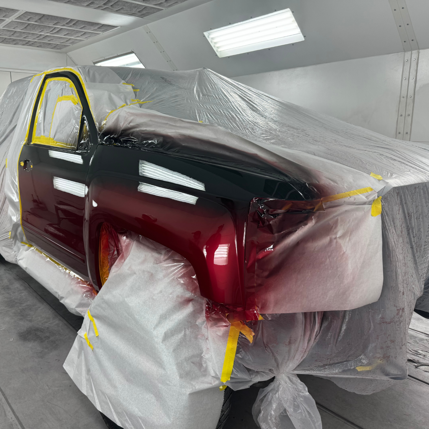Truck cab being painted in a paint booth. Red and black gradient paint job, masked with plastic and yellow tape.