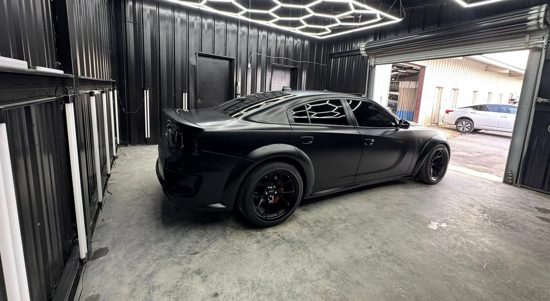 Black car in a garage with honeycomb ceiling lights. The car is matte black and has black wheels.