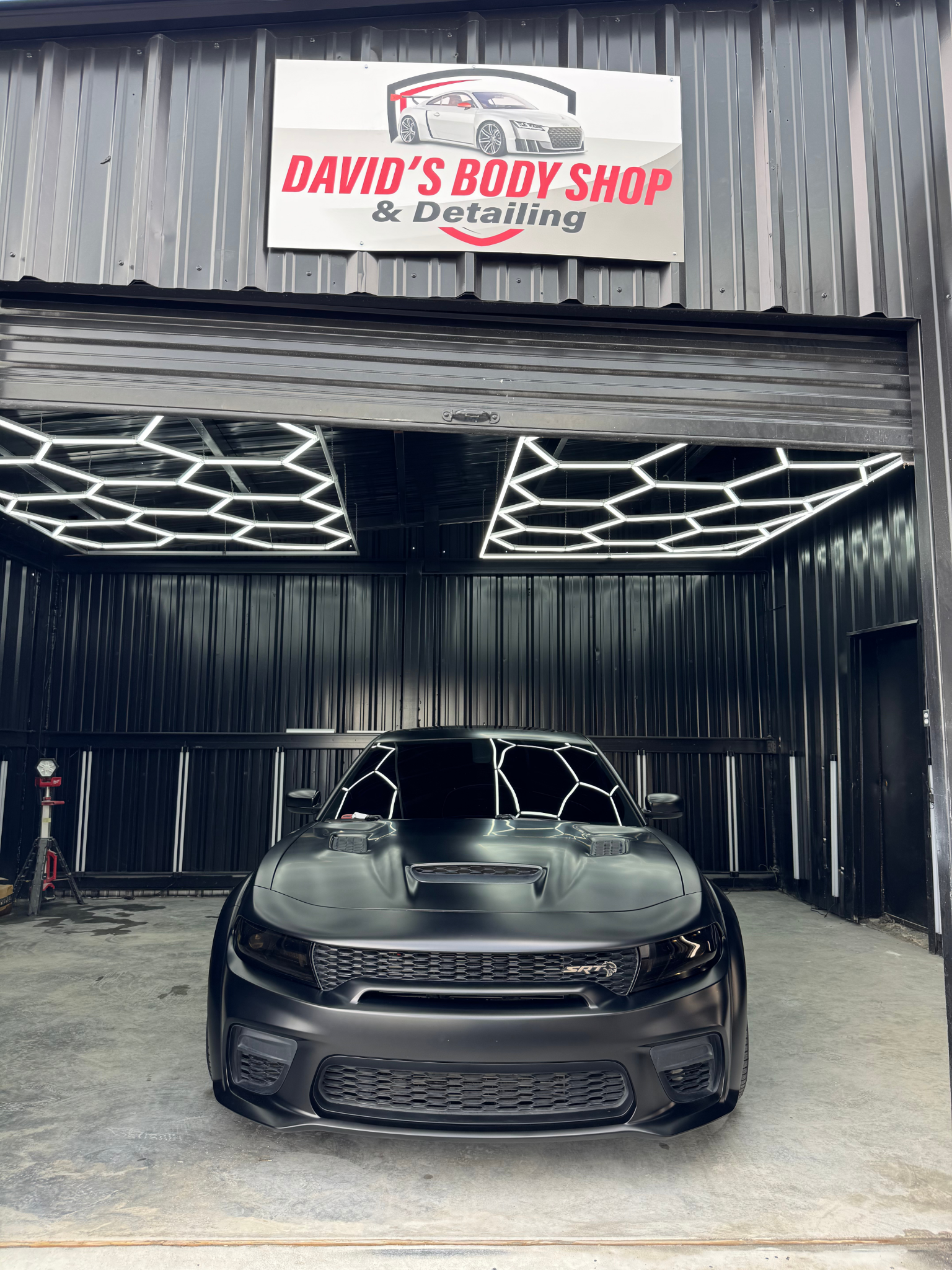Black car inside a body shop with overhead honeycomb lights and shop sign