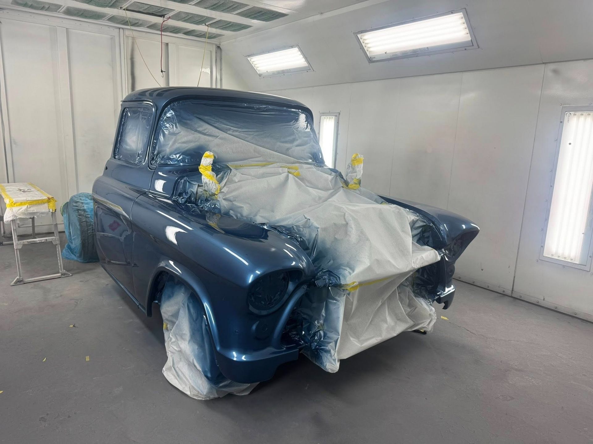 Blue classic truck in a paint booth, partially covered in plastic, ready for paint.
